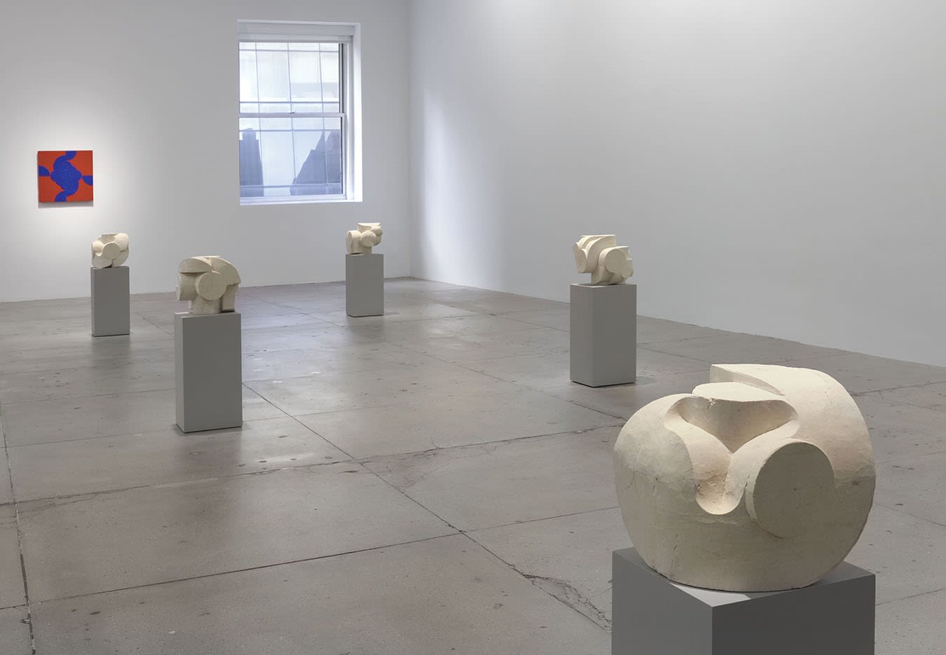 8 small white stone sculptures on pedestals in front of a red abstract painting on a white wall.