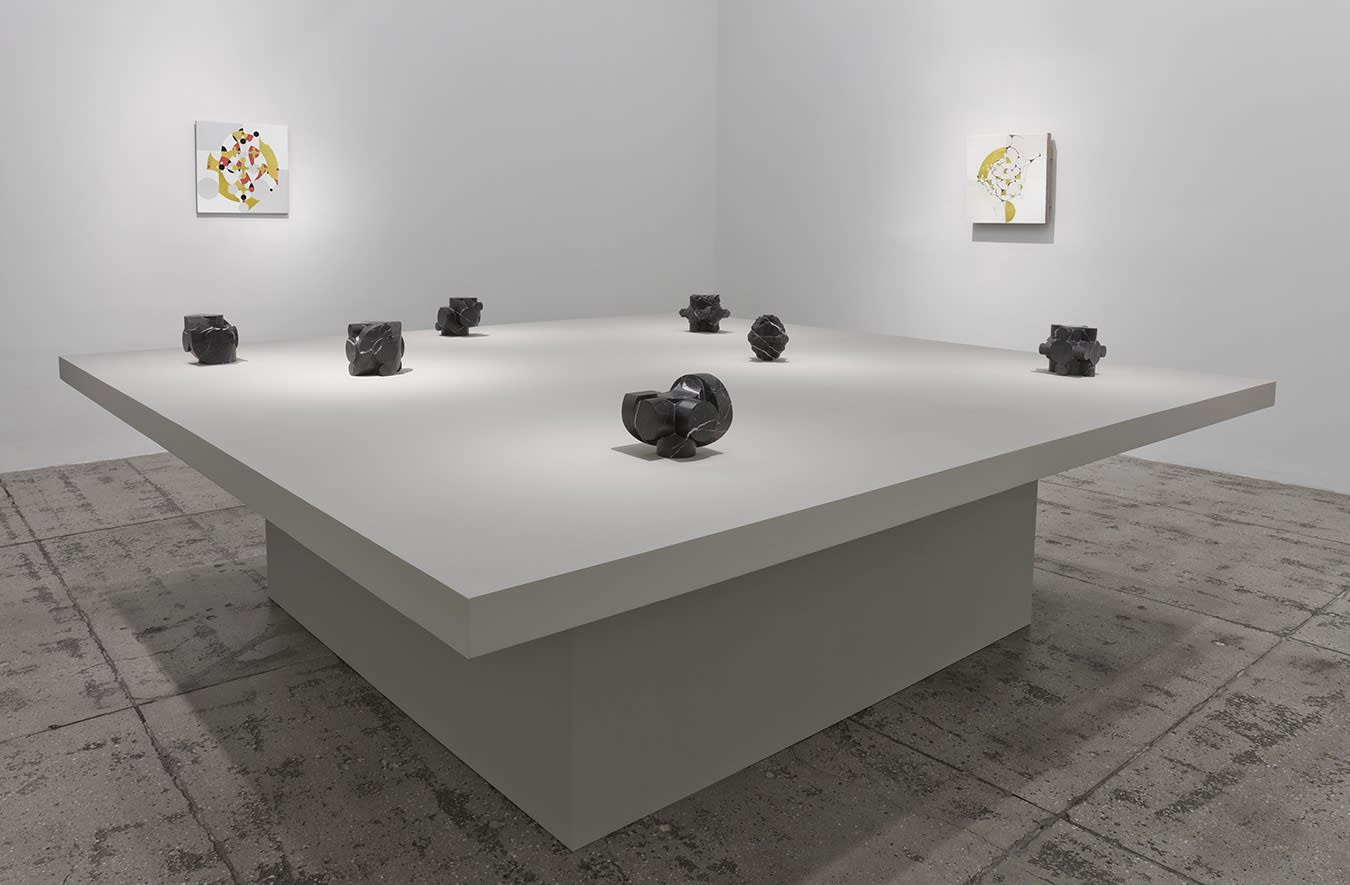7 small stone sculptures on a table in front of two abstract paintings.