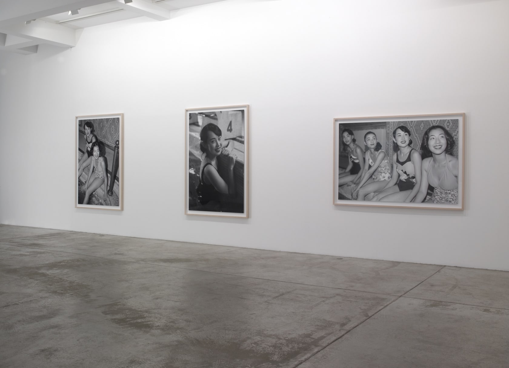Three framed black and white photographs: one of two women sitting on the edge of an indoor pool, another of a single woman in the pool, the final of four women smiling at the camera. 
