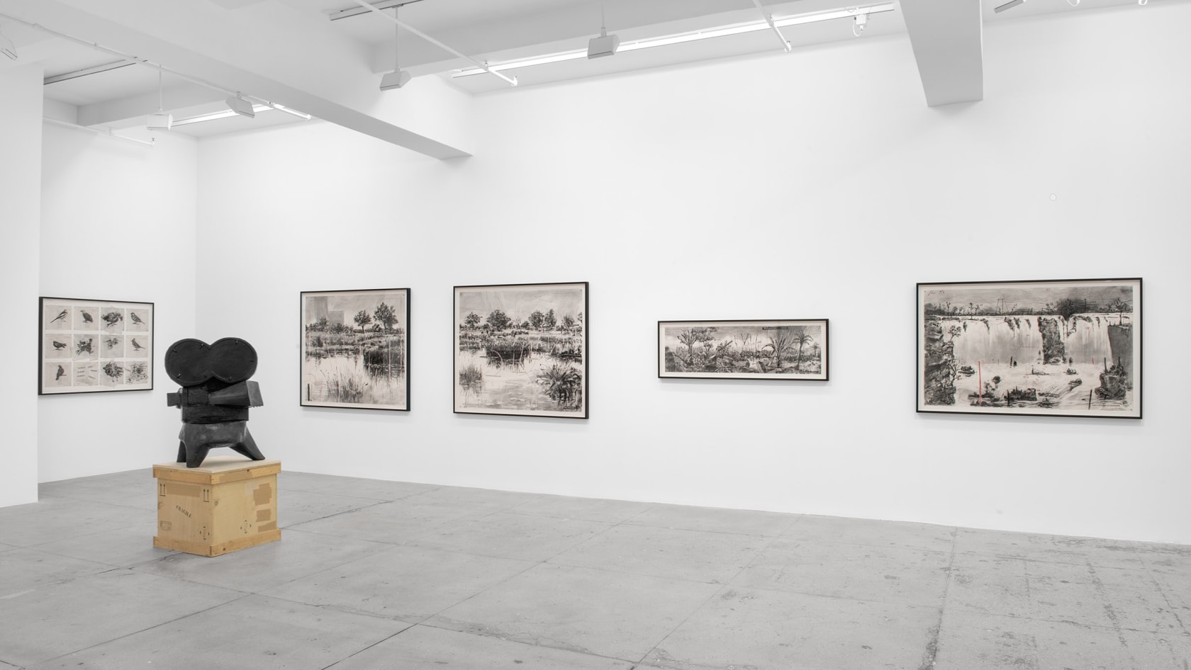 A copper sculpture of an old Hollywood camera stands in front of five graphite drawings of maps, plants and landscapes. 