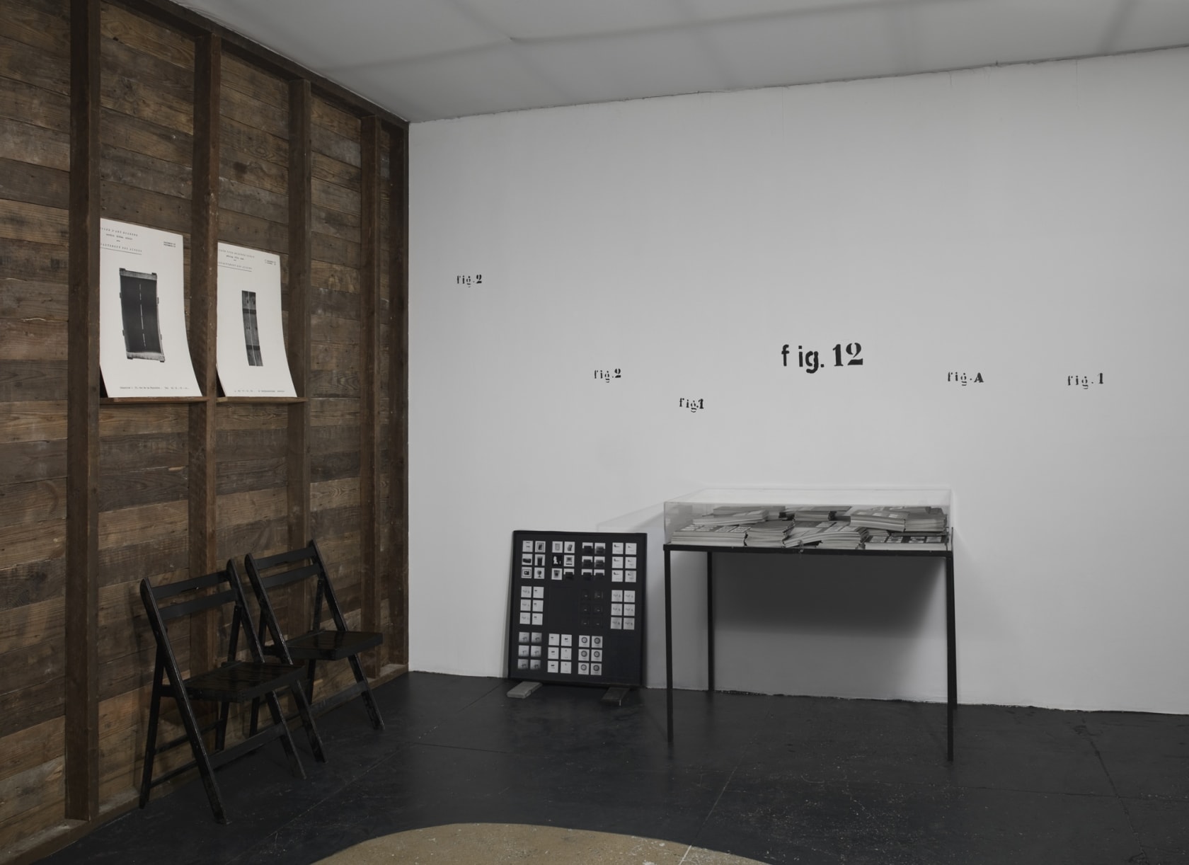 In a room with black floors, a white ceiling, a wooden wall on the left and a white wall straight ahead, there are two chairs by a table. The table is covered in stacks of paper, which is then covered by a glass barrier. Text on the white wall reads: fig.
