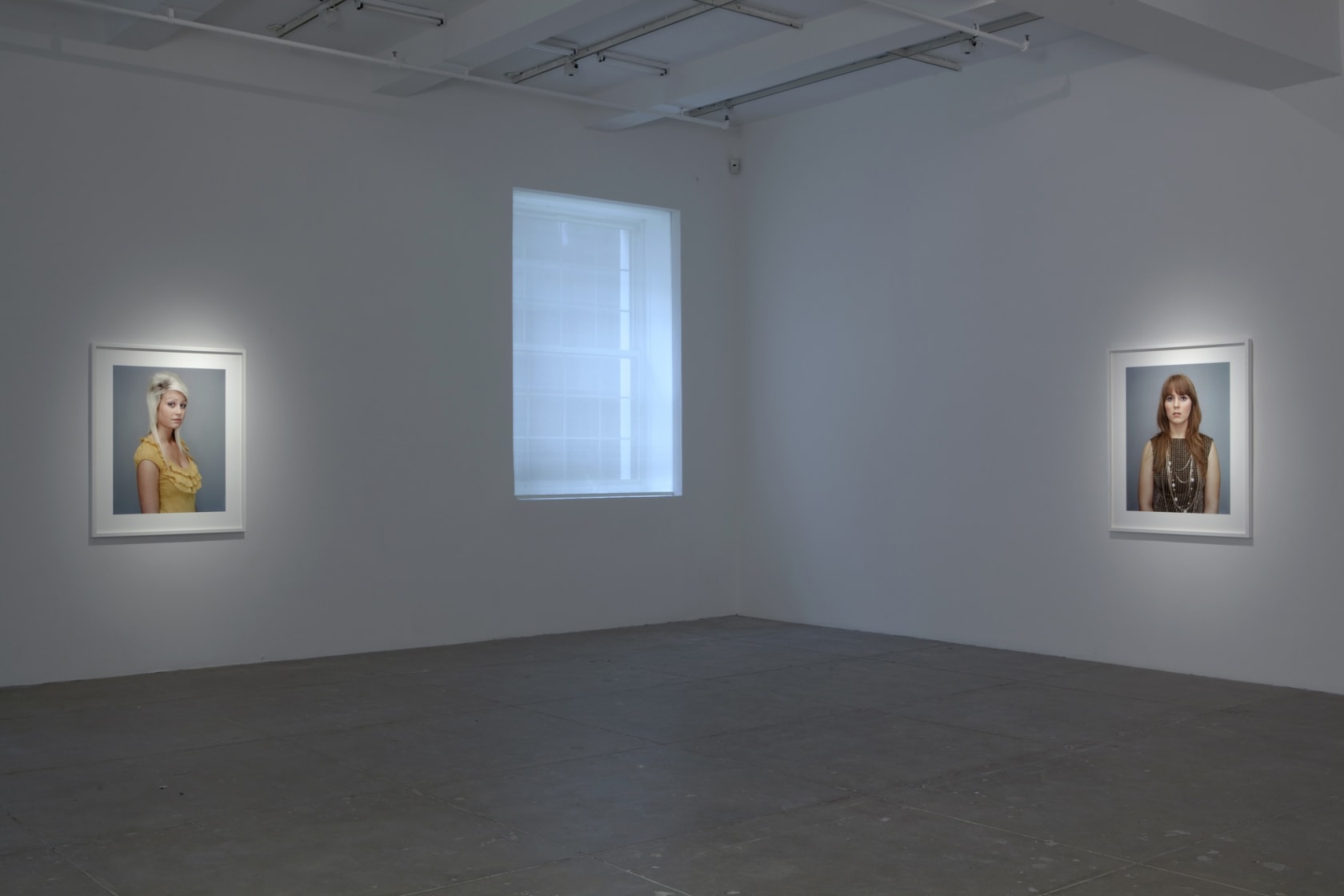 On white walls in a large space, two portraits of young women hang, with a large window in between them. One woman wears a yellow top, the other brown with several silver necklaces.