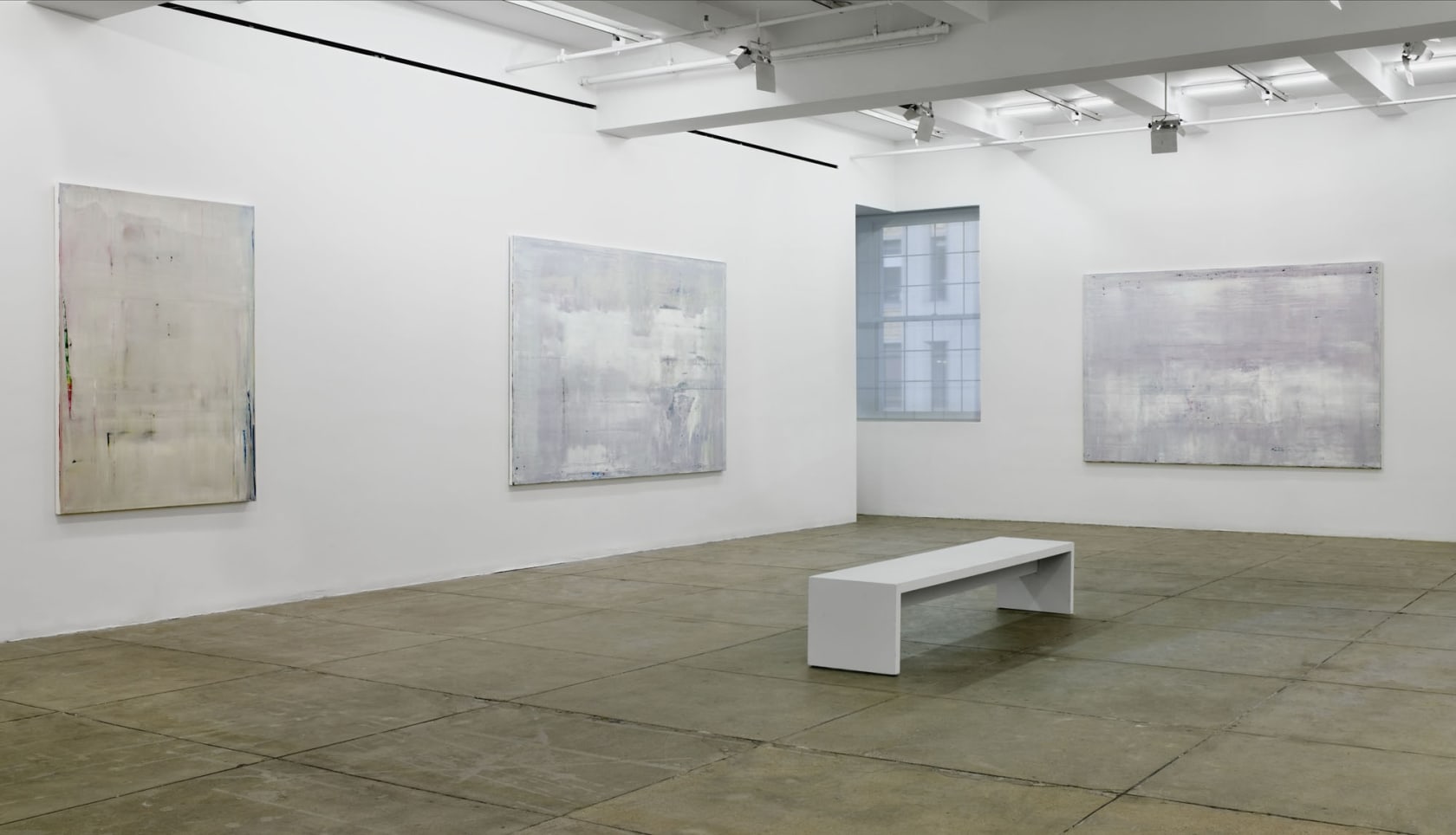 3 large, light grey, abstract paintings hang in the corner of a room. There is a window in the corner and a white bench in the center of the floor. 