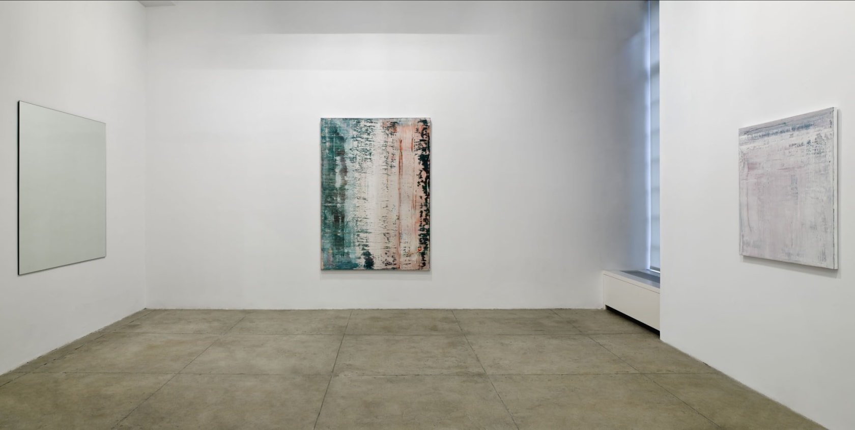 3 abstract paintings hang on 3 walls forming a corner. There is a window on the right. 
