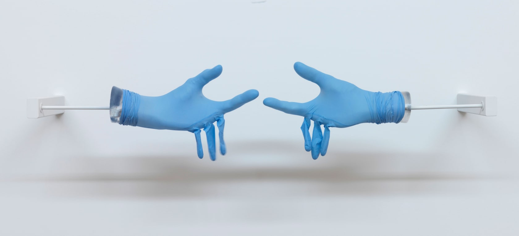 Two blue medical gloves are stretched over plaster hands reaching for each other. The middle, ring and pinky finger are empty and hang limply. 