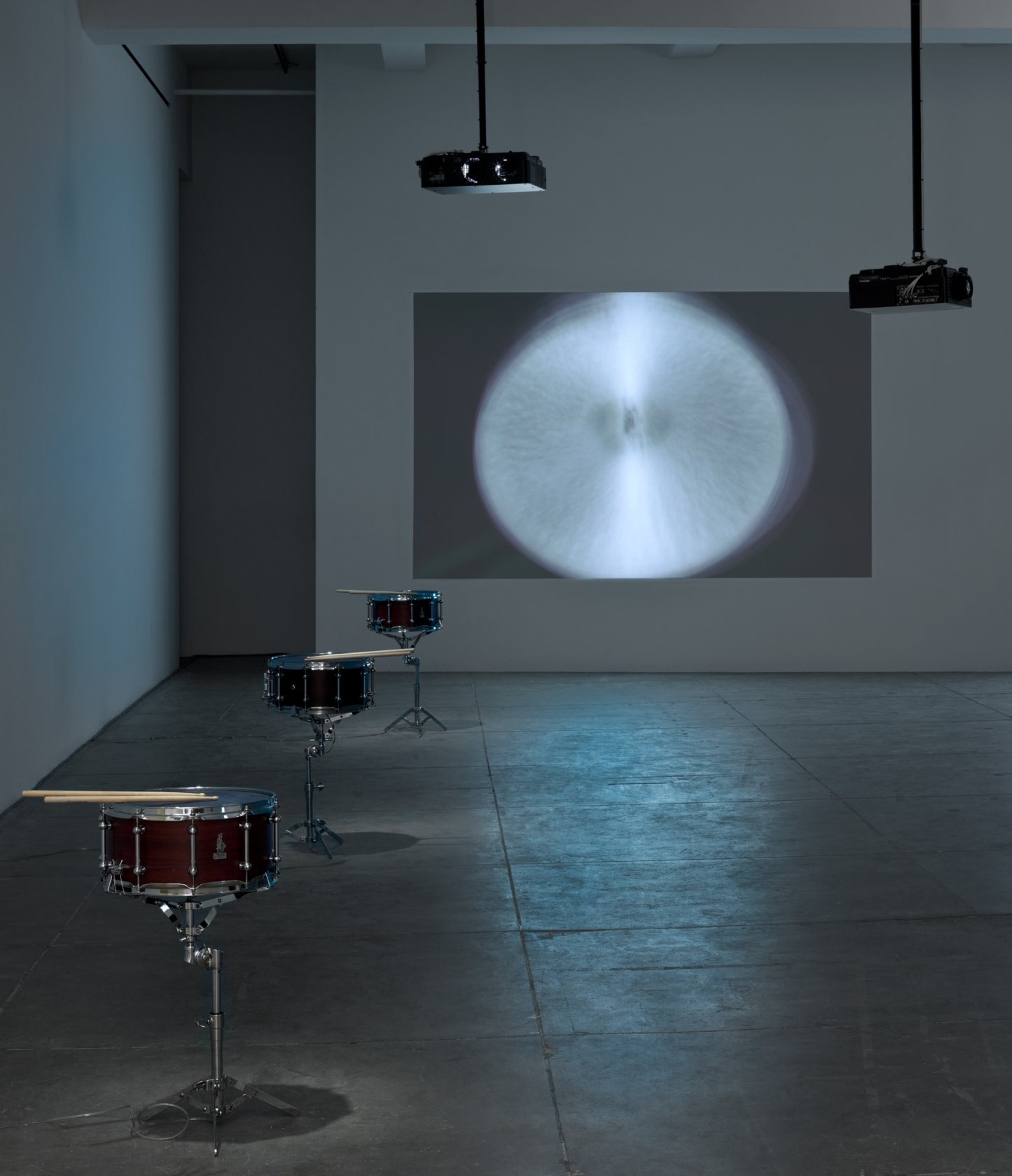 A cymbal with a motion blur is projected onto a wall. 2 black projectors are suspended from the ceiling. 3 snare kits sit in a line on the left side of the room. 