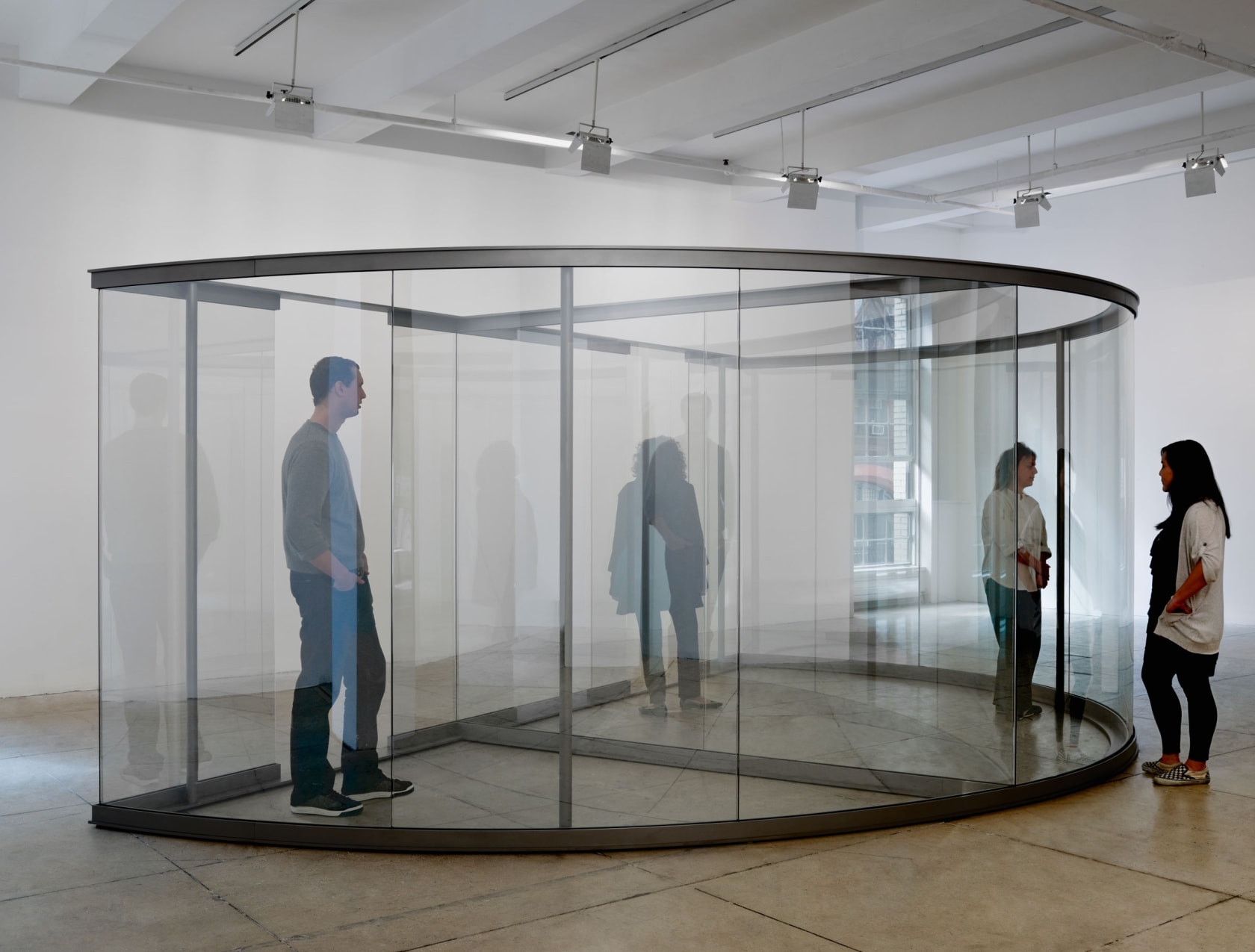 Various people move through a steel-framed half circle with glass panels, distorting their reflection. 