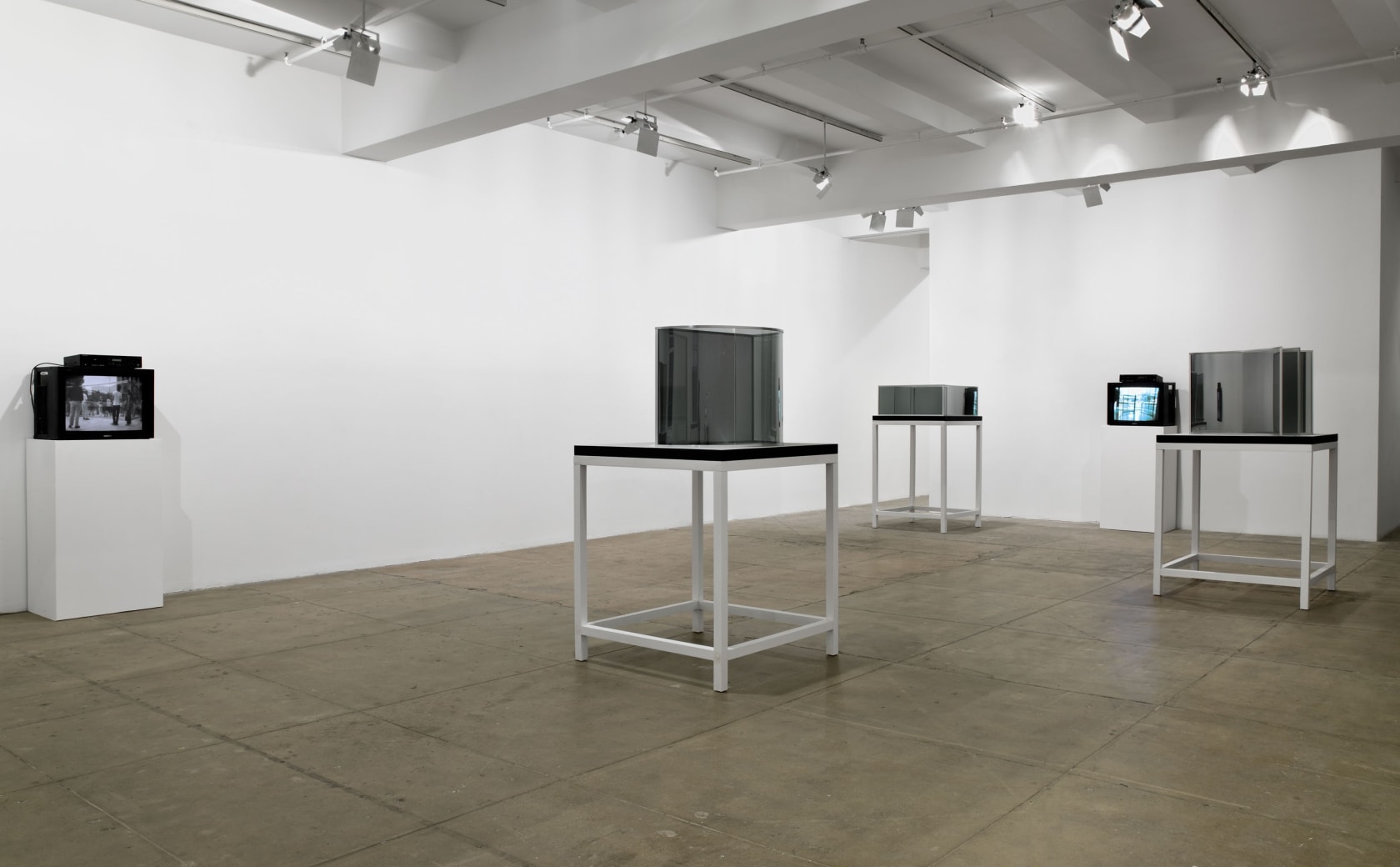 Three glass and mirror sculptures are installed atop wooden tables; two tv's nearby are reflected in the surfaces. 