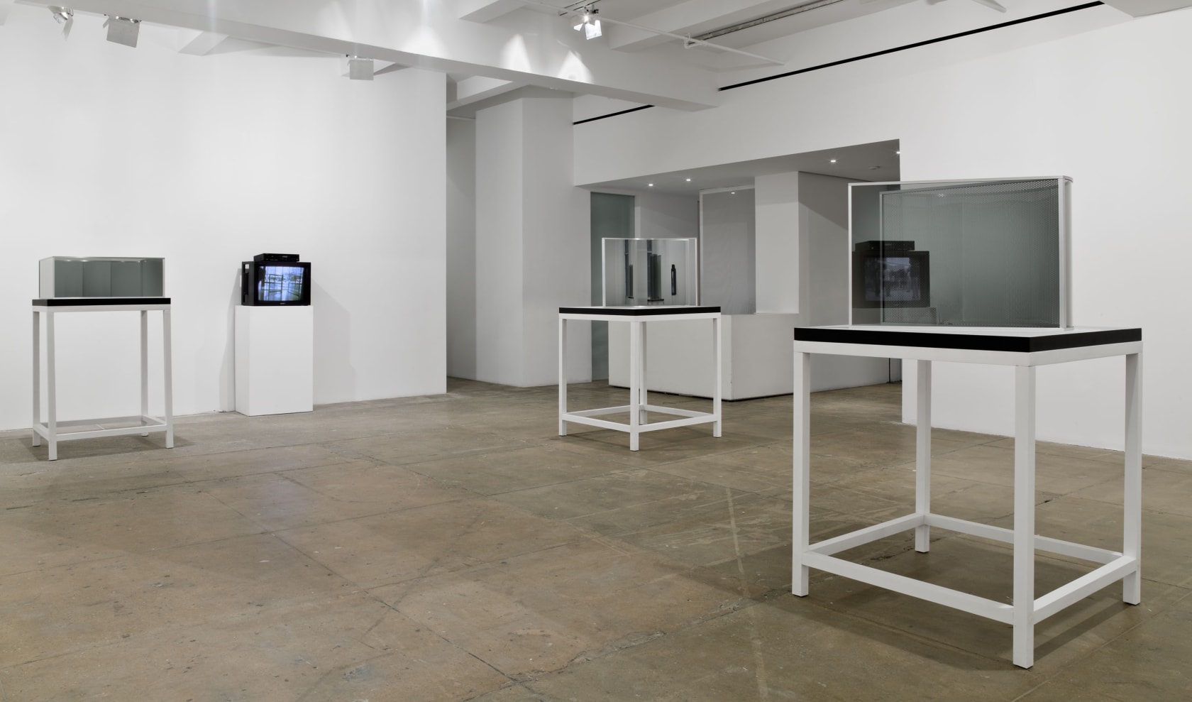 Three glass and mirror sculptures of different shapes are installed atop tables; a tv stands between projecting images. 