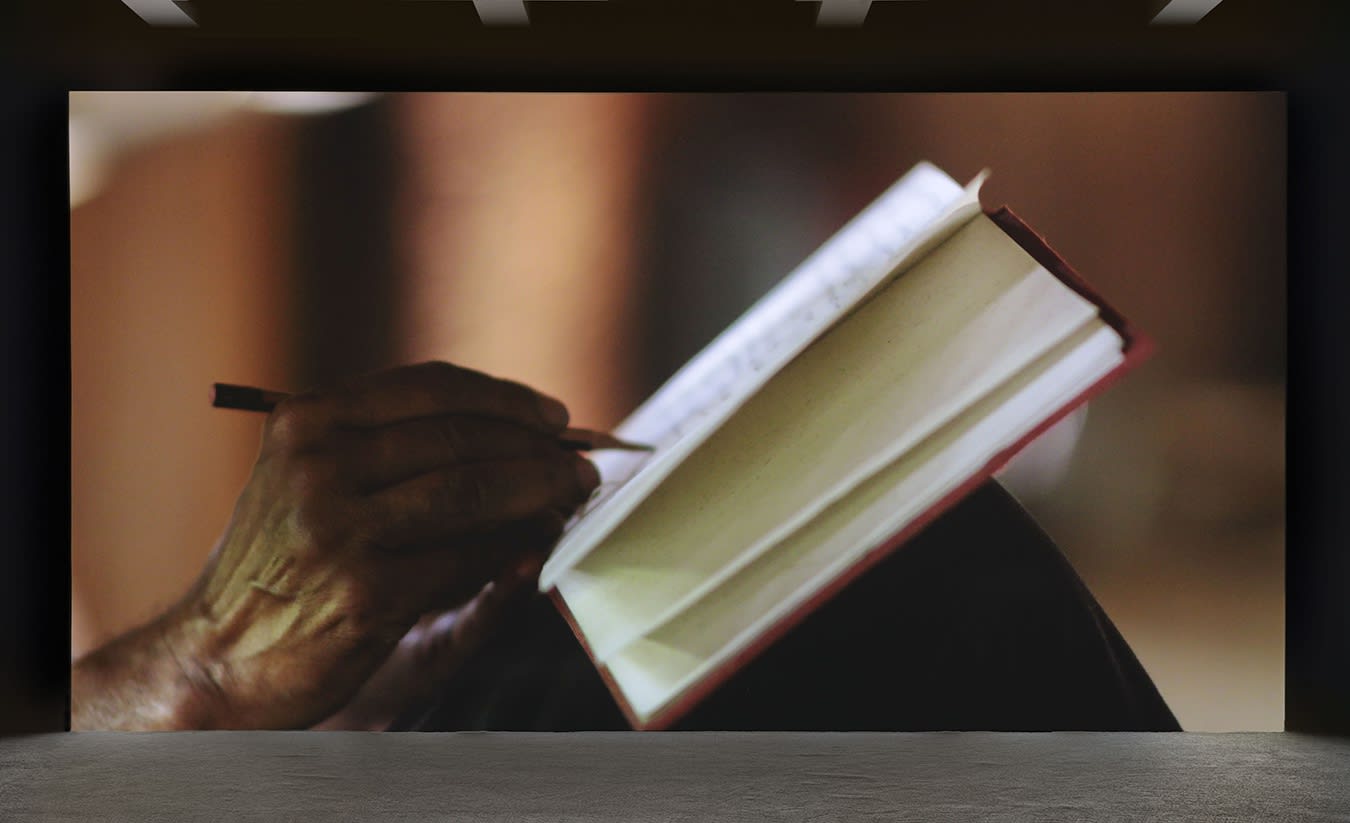 A large projection depicts a close-up of an elderly hands writing in a book with a pencil. 