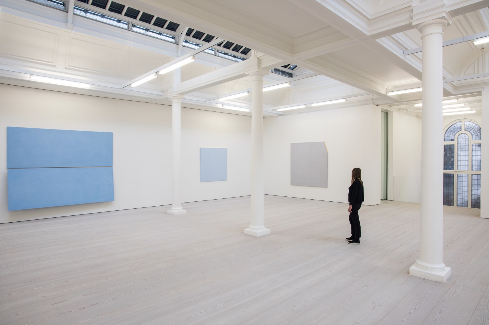 In a large white space with columns, two large light blue paintings hang, with one light grey painting. A woman dressed in black looks at the art.