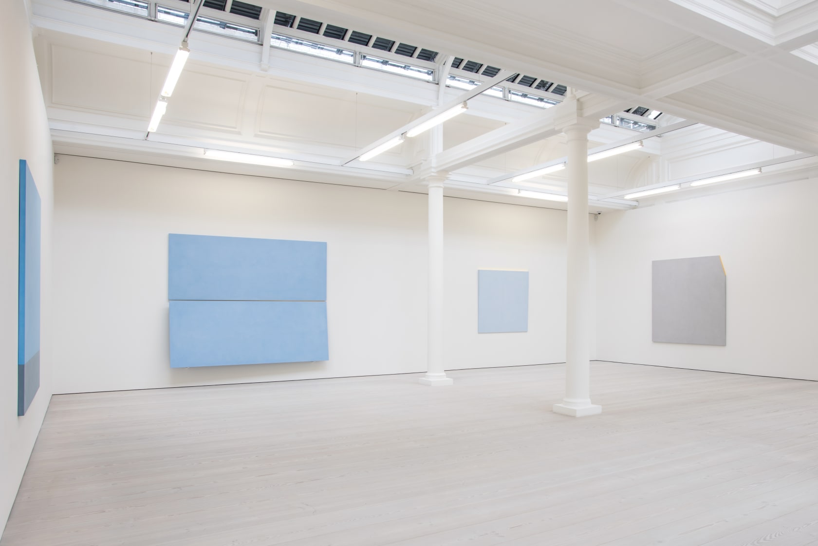 4 blue and gray paintings hang in a large white room. There is a long skylight in the ceiling and 2 white columns. 