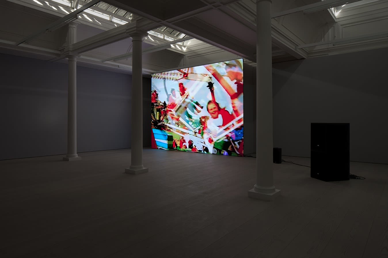 A projection screen stands in a darkened room with 3 columns, a skylight, and 2 black speakers. The projection displays people laughing on an amusement park ride. 