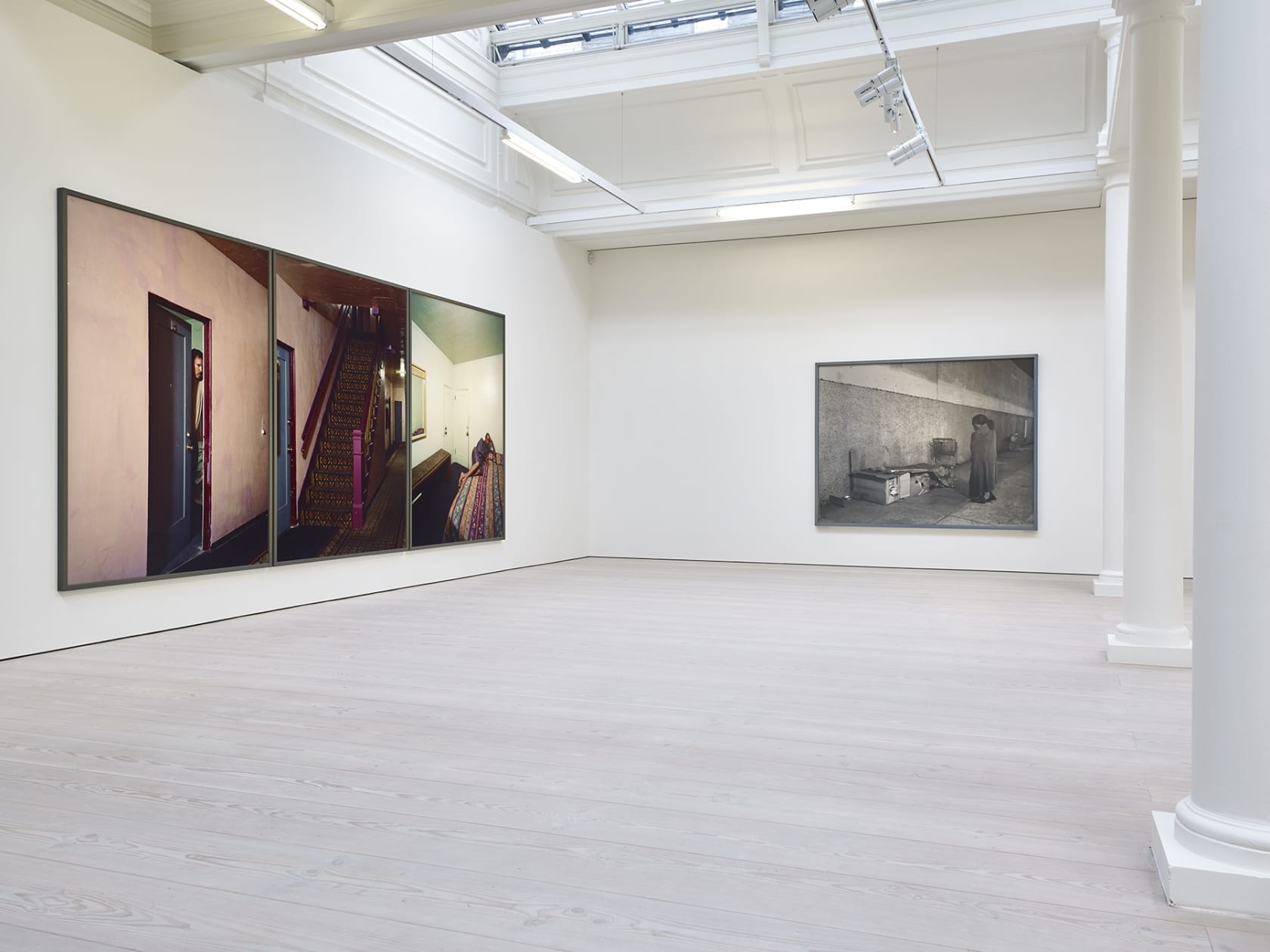 4 large photographs hang in the corner of a white room with skylights. On the left, 3 photos make up a panorama of an interior with a staircase, while on the right is a black and white image of a person wrapped in a blanket on the sidewalk. 