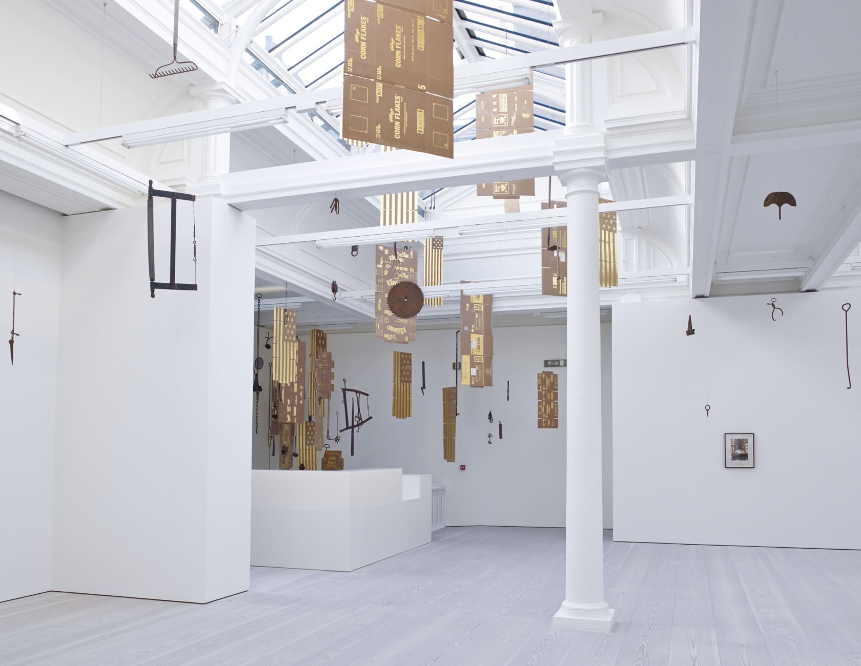 In a large white space with a long glass skylight, sculptures hang from the ceiling - most seem to be American flags, with 13 stars, made out of gold painted cardboard. Another is an unfolded Corn Flakes box, made from the same material.
