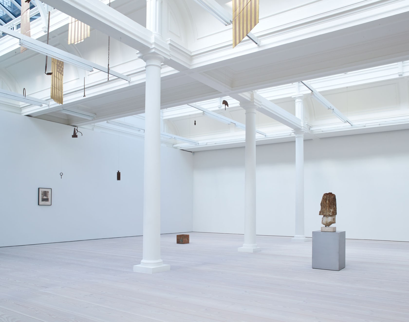 In a large white space with a long glass skylight, sculptures hang from the ceiling - most seem to be American flags, with 13 stars, made out of gold painted cardboard. A small cardboard box sits on the floor as well.