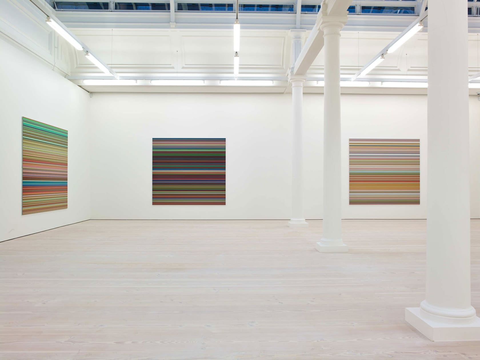 In a large white space with columns and skylights, three paintings hang, all square and about 6 by 6 feet. They are all made up of hundreds of perfect horizontal strips of various colors.