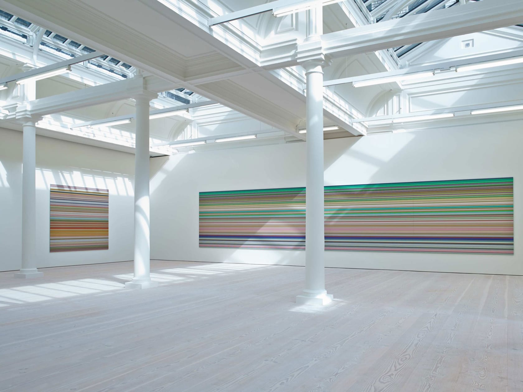 In a large white space with columns and skylights, an enormous painting hangs, light from the skylight shining on it. It is made up of hundreds of perfect horizontal strips of various colors, mostly light green in hue, with a great deal of reds and blues.