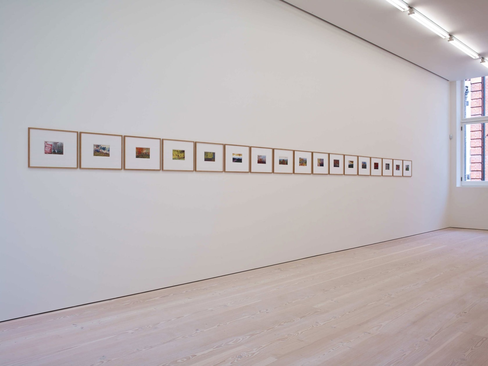 On a long white wall, about 15 paintings hang, all very small, and bordered by large white frames, further bordered by wood. The paintings are all done over prints of film photographs.