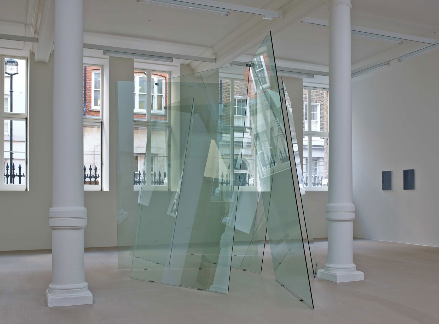 In a large white space with columns and many windows, several glass planes lean on each other unevenly, making a large sculpture.