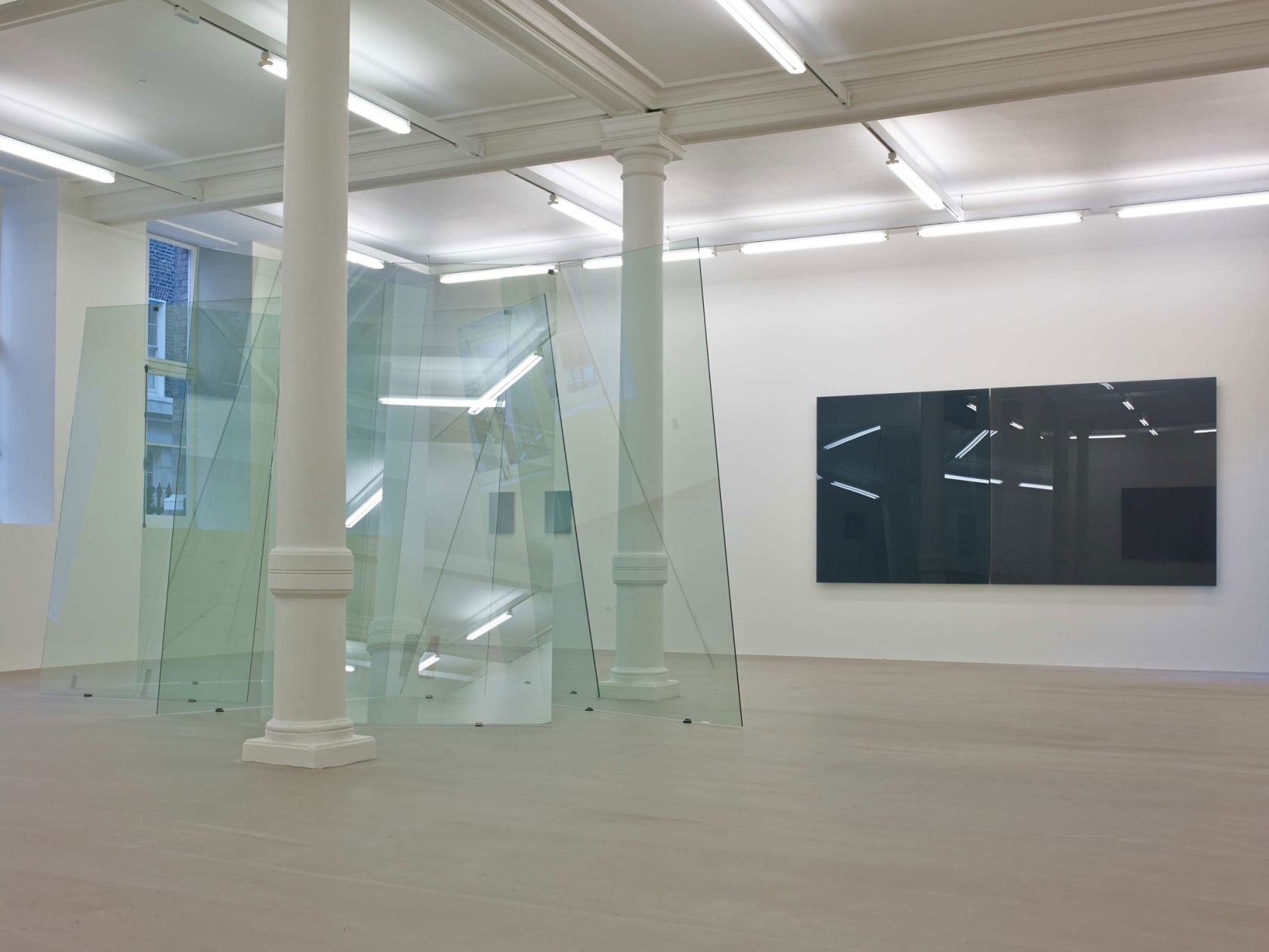In a large white space with columns and many windows, several glass planes lean on each other unevenly, making a large sculpture. Behind it on a wall, two large square glass panes hang, one black and one grey.