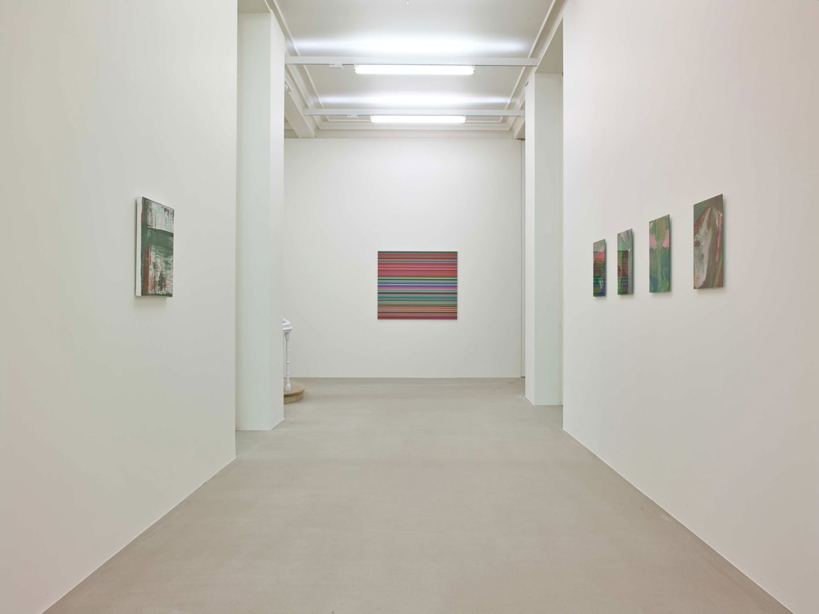 On a white wall down a hallway is a square painting, made up of hundreds of perfect horizontal strips of various colors. On both walls of the hallway hang small abstract paintings (1 right, 4 left), mostly green in hue.