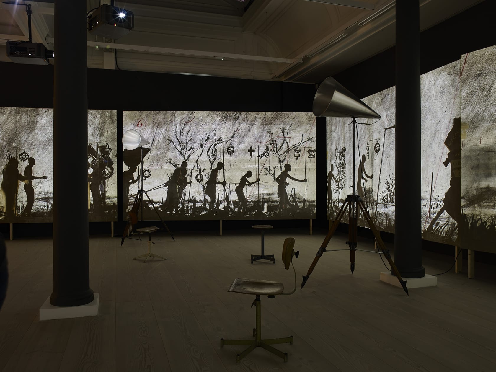 In a large dark space with columns, stools, and large objects resembling megaphones, several large video screens create a panoramic film, showing several characters, both people and sculptural representations of people's heads, walking through a marsh.