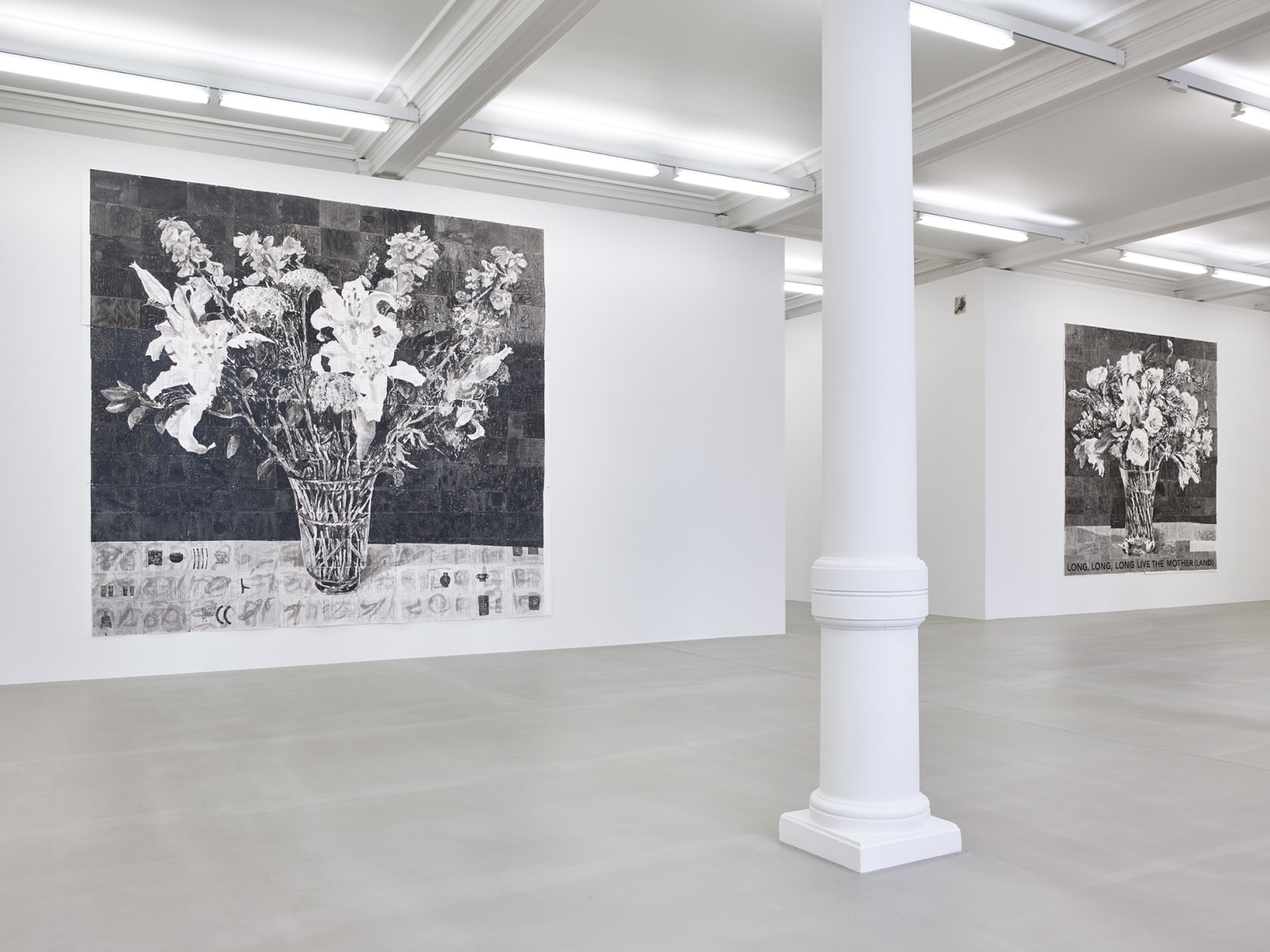 In a large white space with columns, two large black and white paintings of flowers in a pot hang on walls opposite sides of a hallway. One on the left reads, at the bottom: LONG, LONG, LONG, LIVE THE MOTHER(LAND)