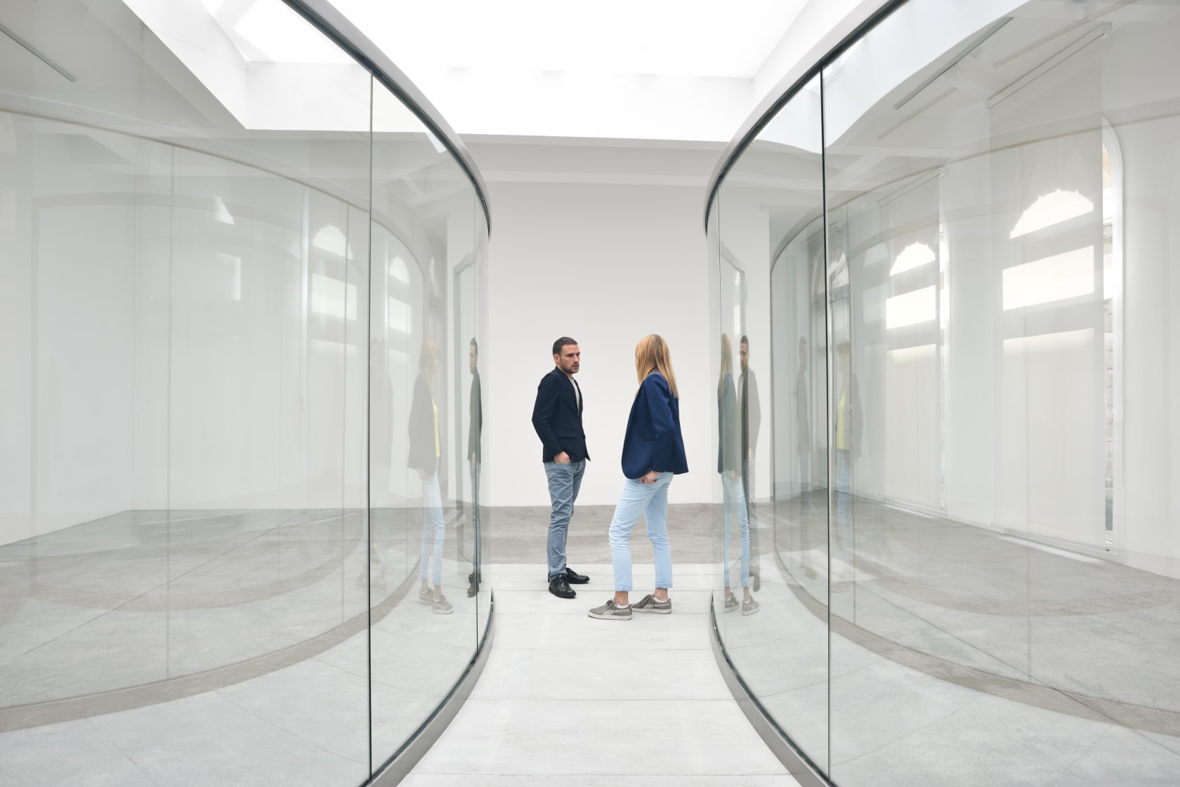 Gallery installation: two people stand in-between two curved glass walls.