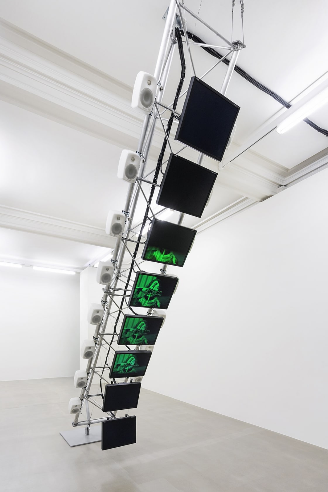 8 flatscreen monitors are displayed vertically on a metal armature in the center of a white gallery space.