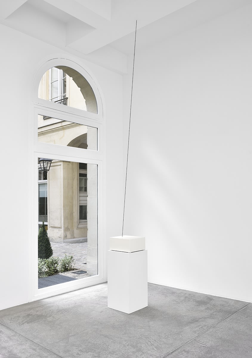 A pedestal sits in front of a window in a well lit gallery. A dark line is suspended from the ceiling.