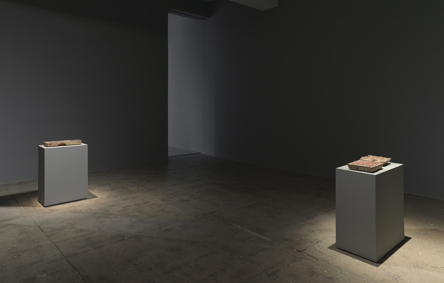 2 grey pedestals spotlit in a grey room displaying flat objects.
