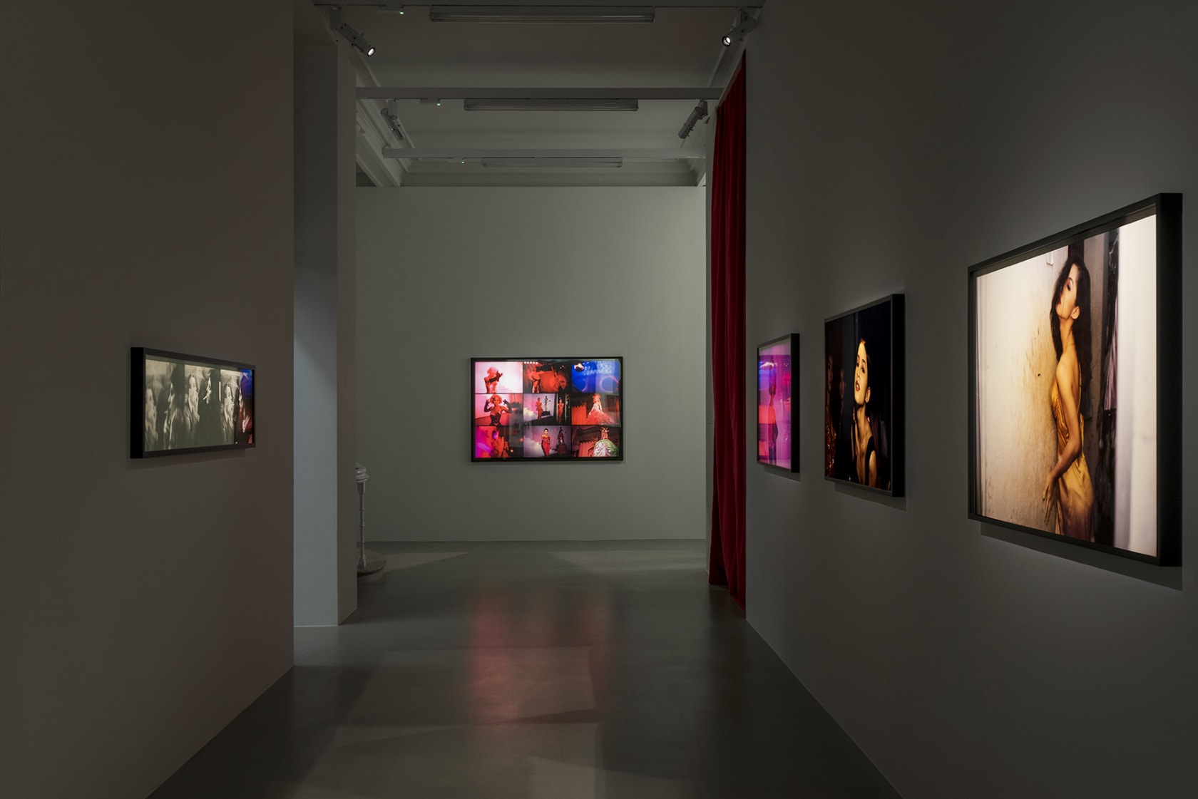 5 spotlit framed photographs on a grey wall with a red curtain leading to another room on the right. 