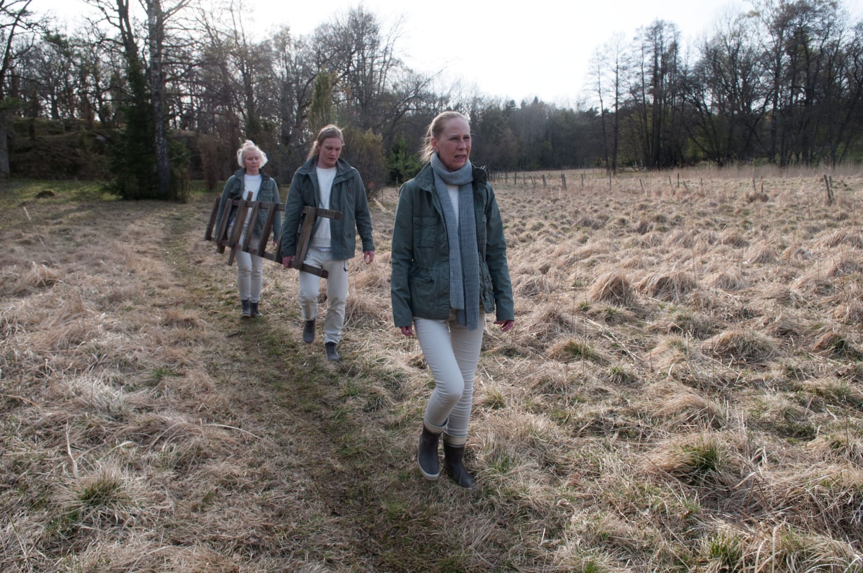 Image of three people in matching green jackets and tan pants walk in a line through a field. Two carry a ladder.