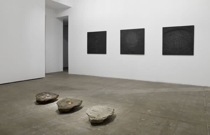 Gallery view. Three flat-topped rocks are parallel to three textured black paintings mounted on the wall.
