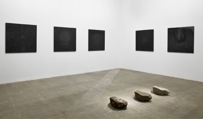 multiple black textured paintings mounted in gallery. Three oval shaped rocks sit facing the far wall in the middle of the floor.