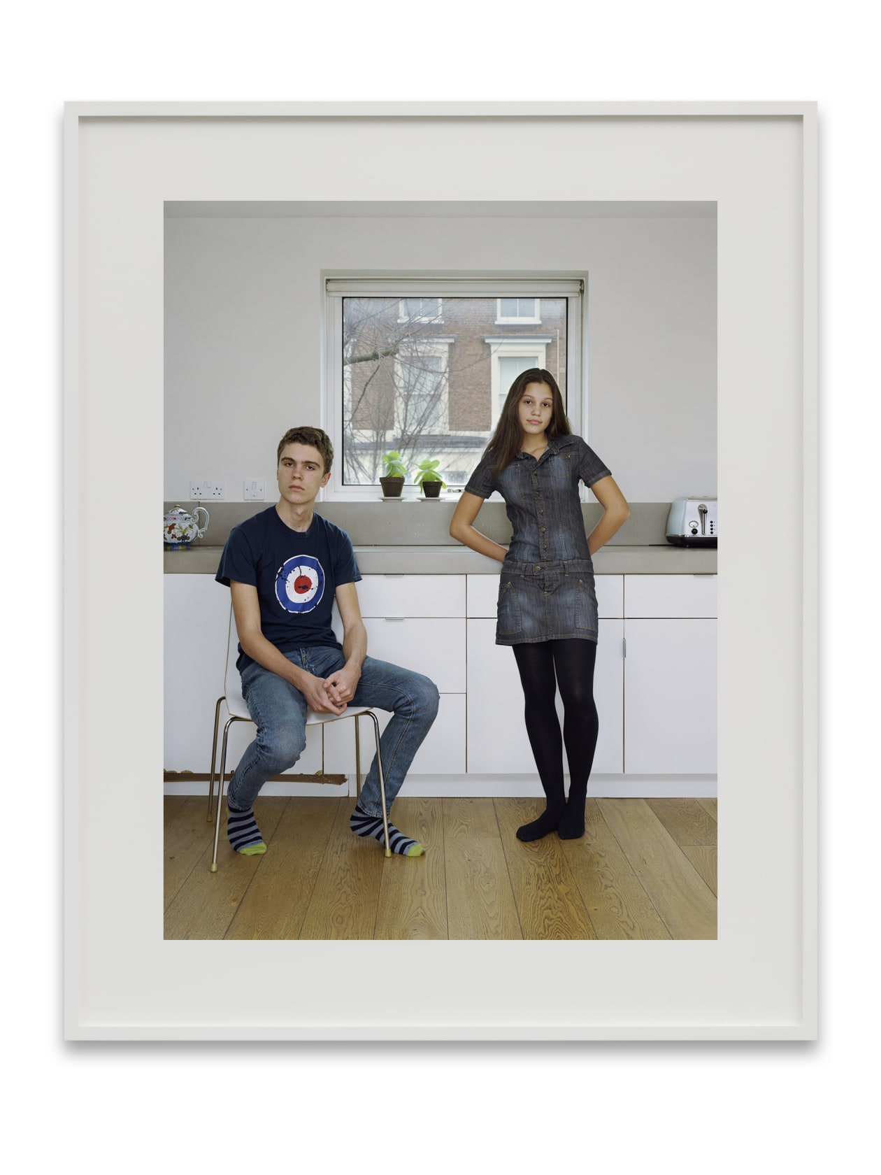 A Rineke Dijkstra portrait: 2 teenage siblings, a boy and a girl, in a white kitchen. 