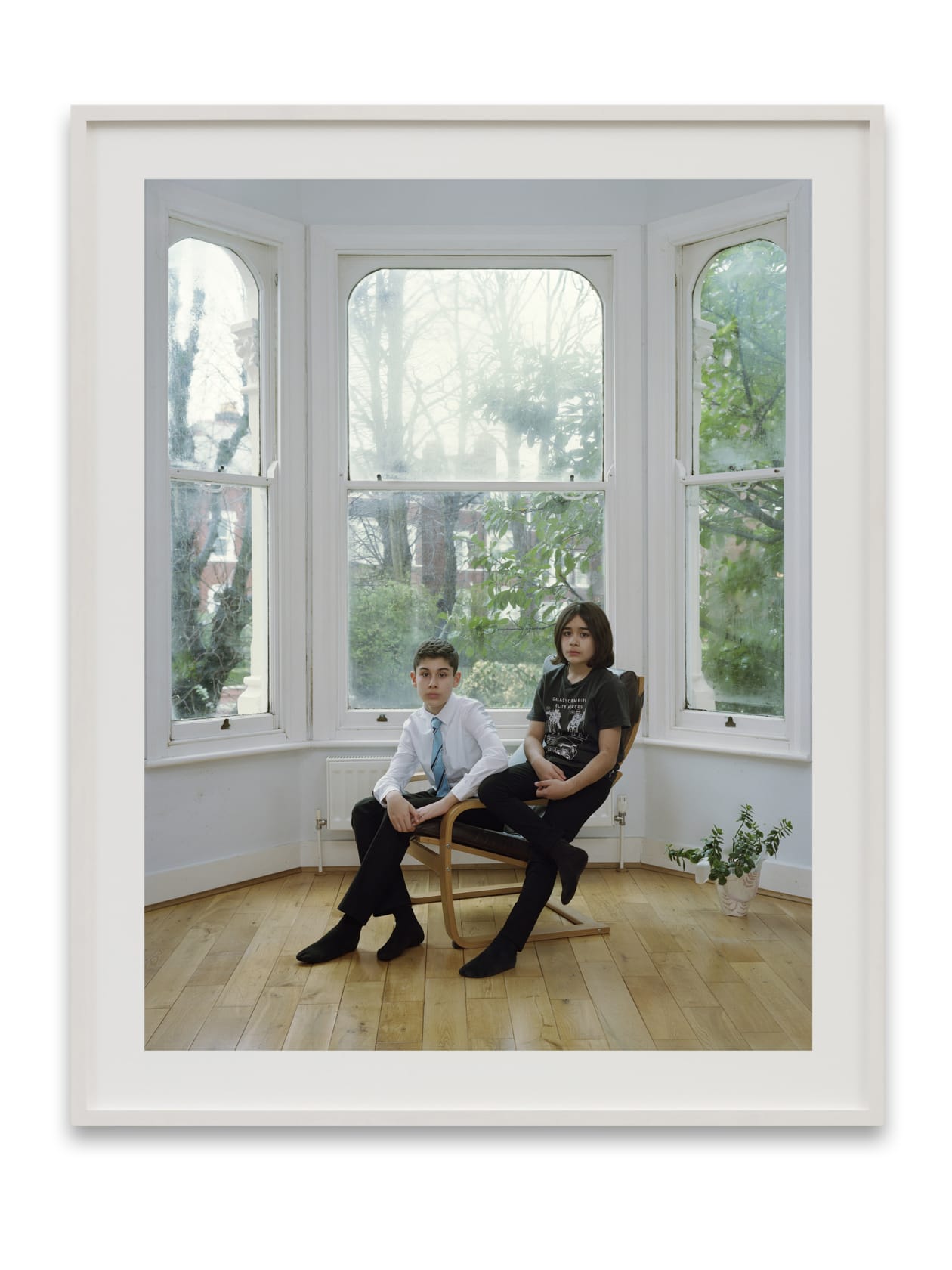 A Rineke Dijkstra portrait: 2 boys, one in a dress shirt and tie, the other in a t-shirt, sit in front of a bay window. 
