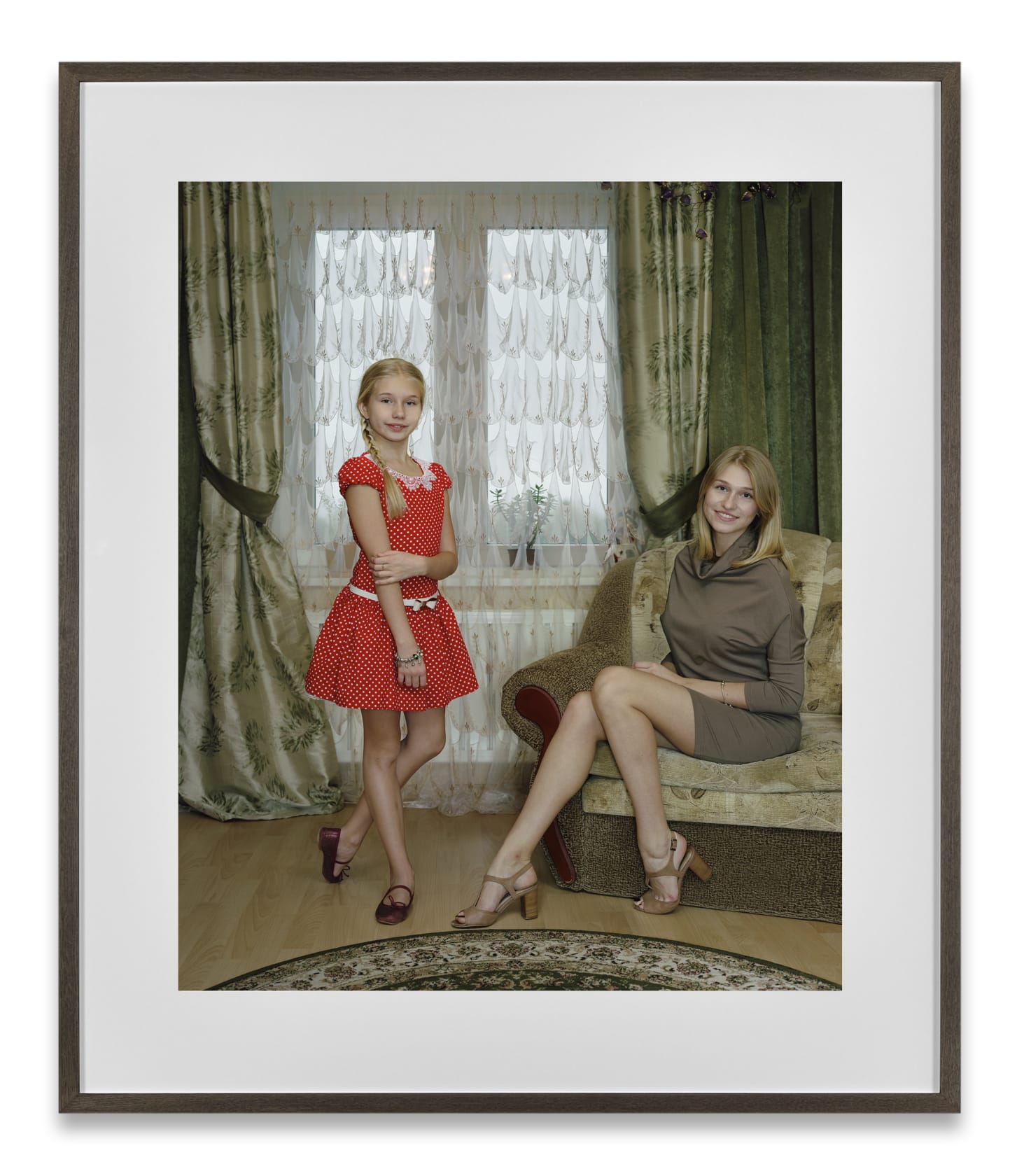 A framed photograph by Rineke Dijkstra of a girl in a red dress and a woman in a beige dress in an elegant living room. 