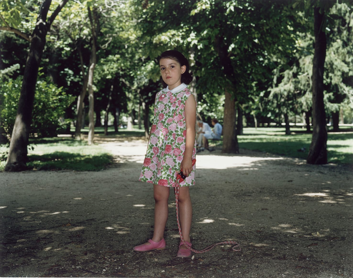 A Rineke Dijkstra photograph: A girl in a floral dress holding a jumprope stands in the center of a tree-lined path in a sunny park. 