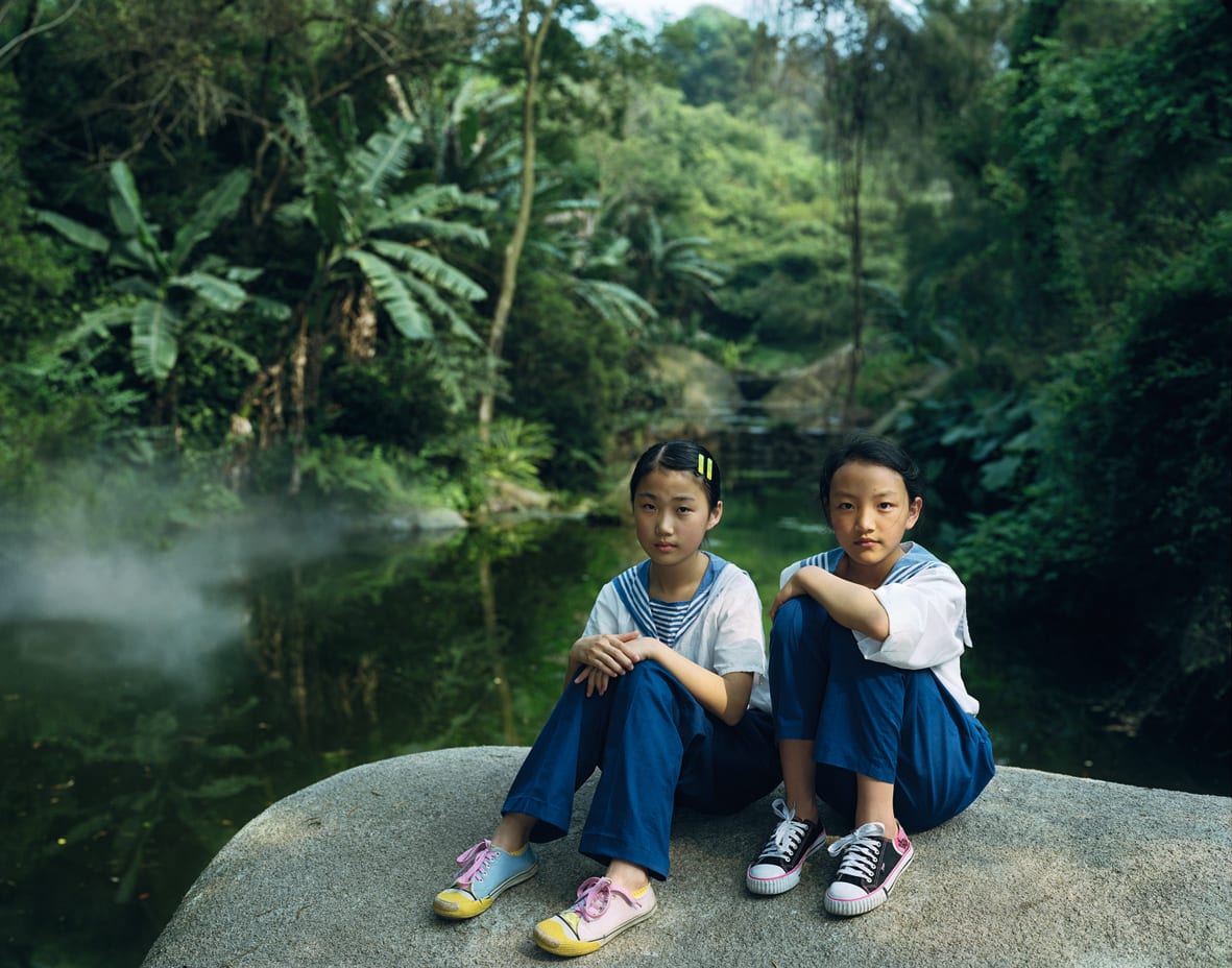 A Rineke Dijkstra photograph: 2 girls in matching uniforms sit on a rock in a lush, green park.