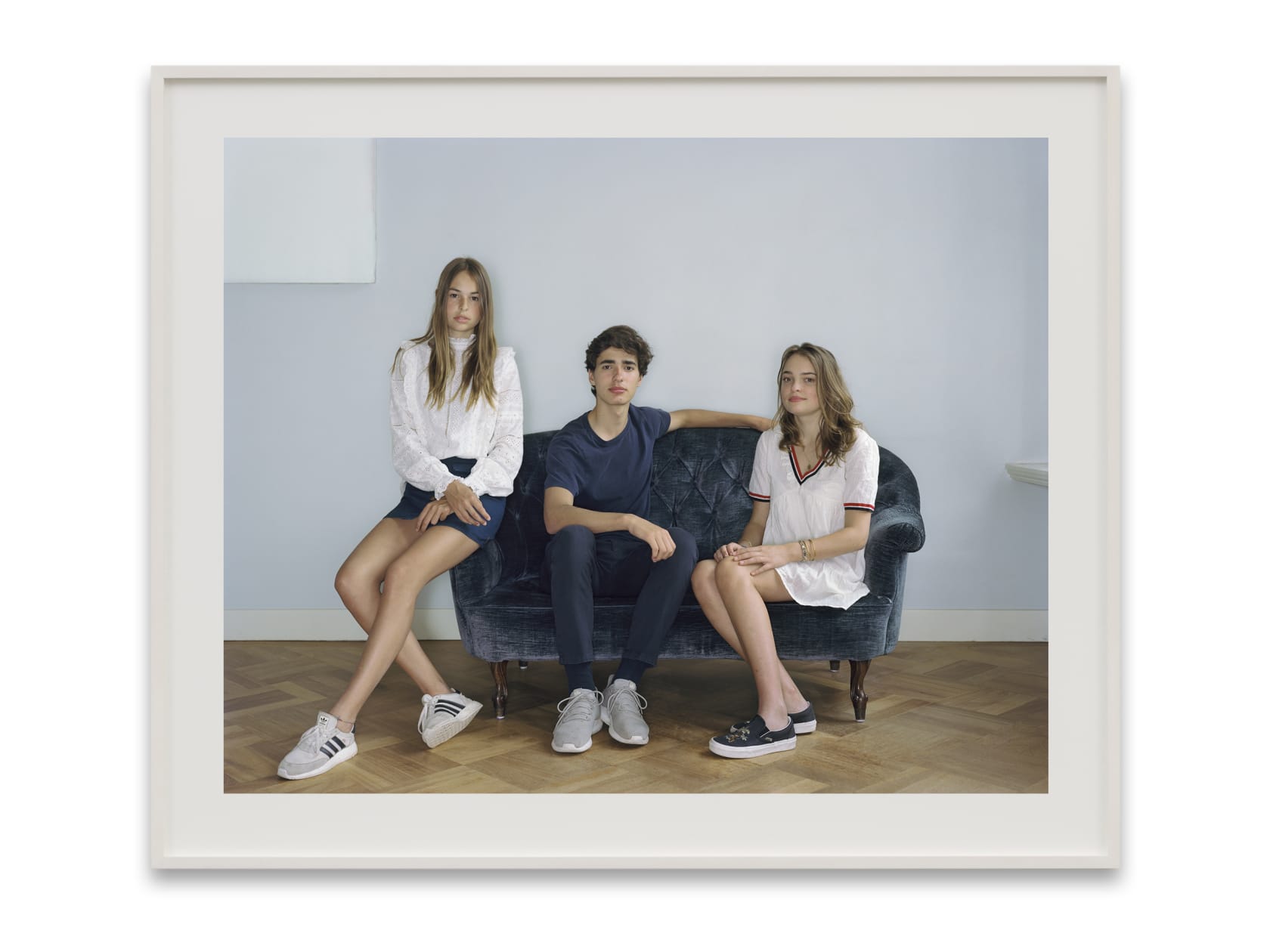 A Rineke Dijkstra portrait: 3 siblings, a boy in between 2 girls, sit on a navy blue velvet sofa.