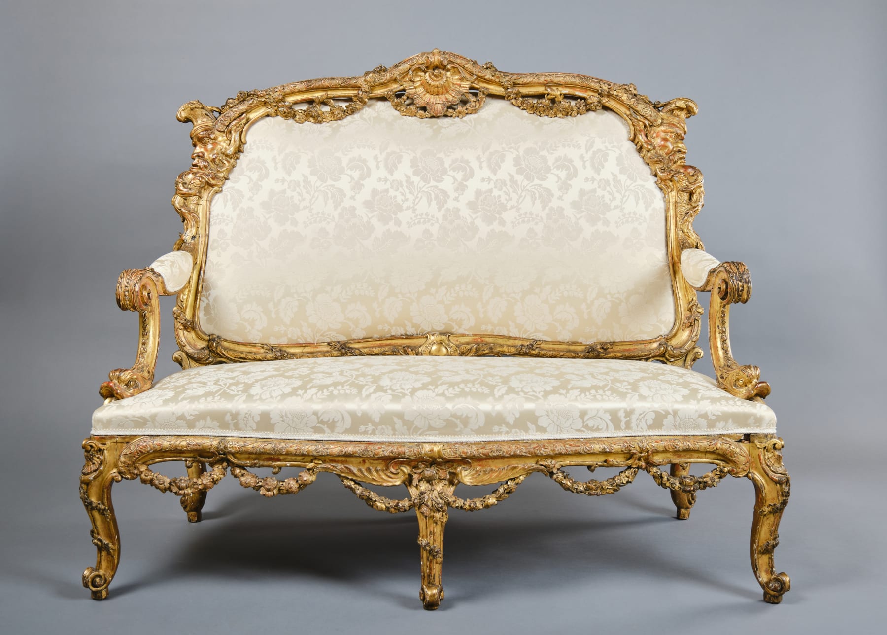 CARVED AND GILDED WOOD SOFA, FRANCE, 18th CENTURY, two-seater, mixtilinear layout and entirely traversed by relief carvings, with floral festoons, garlands, manly masks in the back uprights and stylized dolphins in the armrests with curly finials, upholstered in modern fabric with classical decoration, the back removable