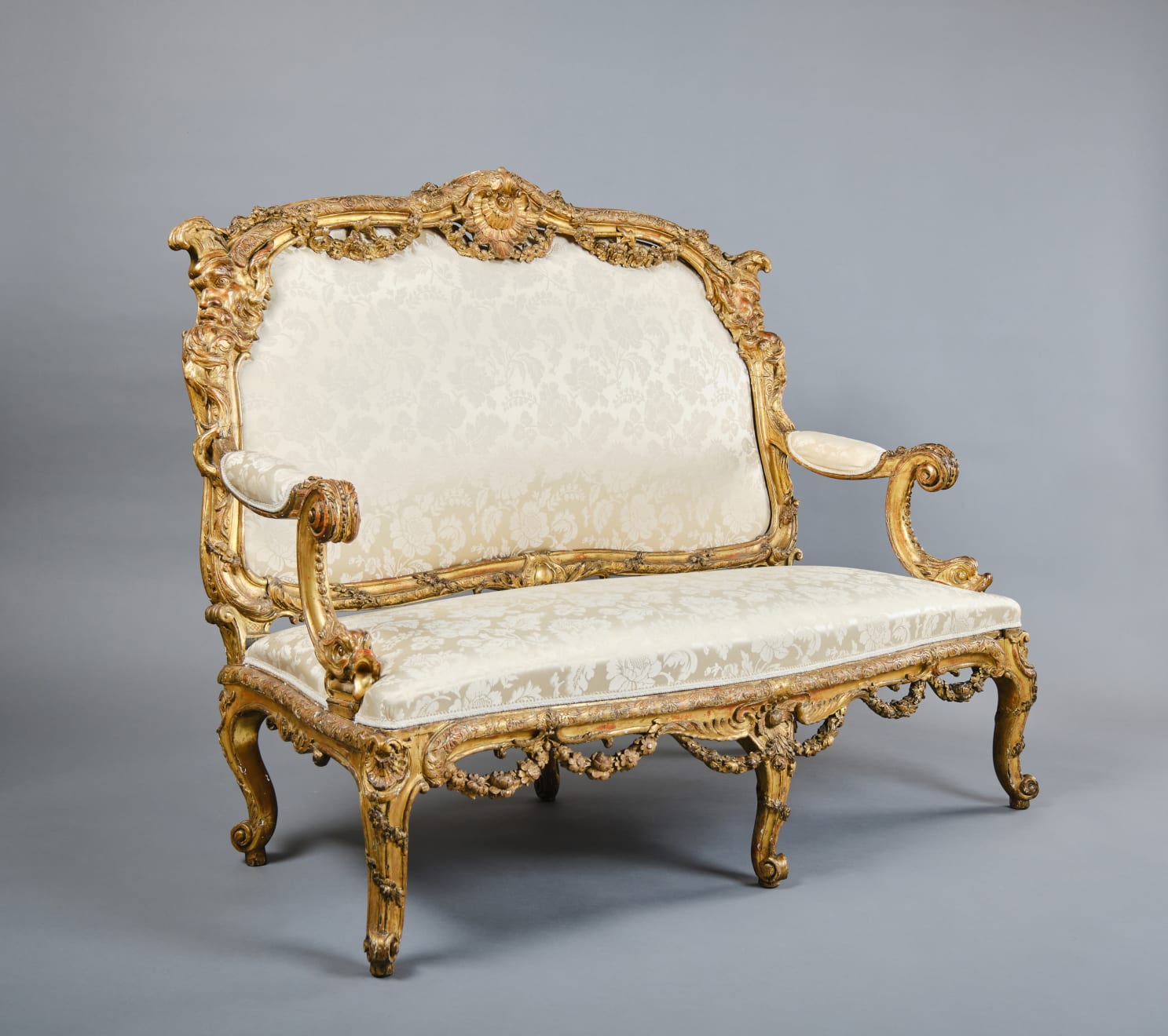 CARVED AND GILDED WOOD SOFA, FRANCE, 18th CENTURY, two-seater, mixtilinear layout and entirely traversed by relief carvings, with floral festoons, garlands, manly masks in the back uprights and stylized dolphins in the armrests with curly finials, upholstered in modern fabric with classical decoration, the back removable