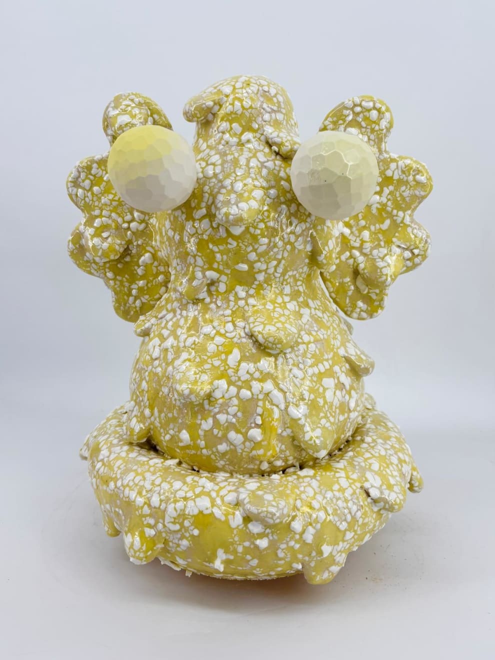 Biomorphic ceramic sculpture titled “Trunkless Baby Elephant” by Kartini Thomas, featuring two pale yellow eyes and a yellow-speckled porcelain surface. Exhibited by Galerie REVEL.