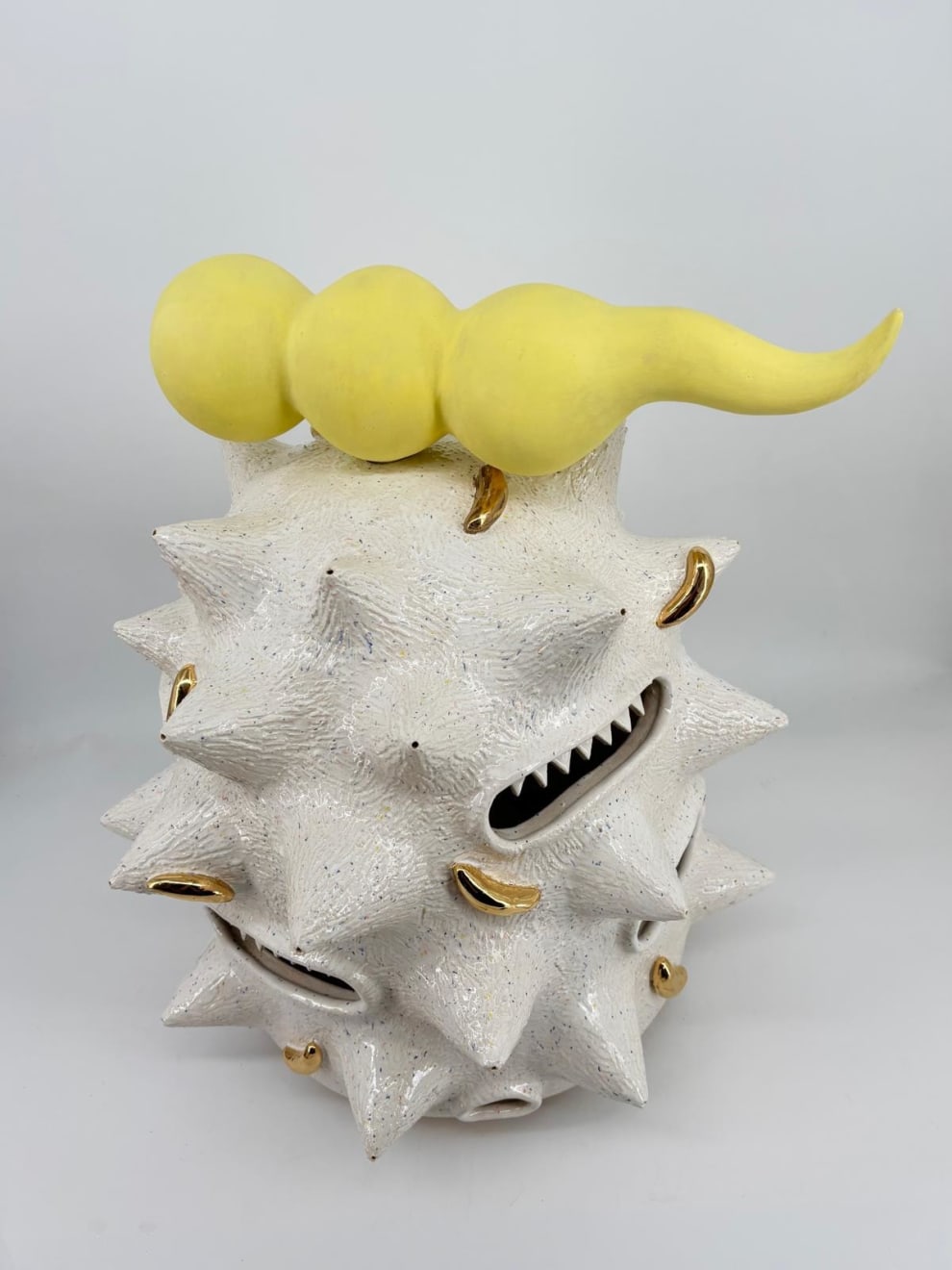 Ceramic sculpture “Fat white spooter” by Kartini Thomas, side view showing spiked white surface with golden details and gaping mouths.