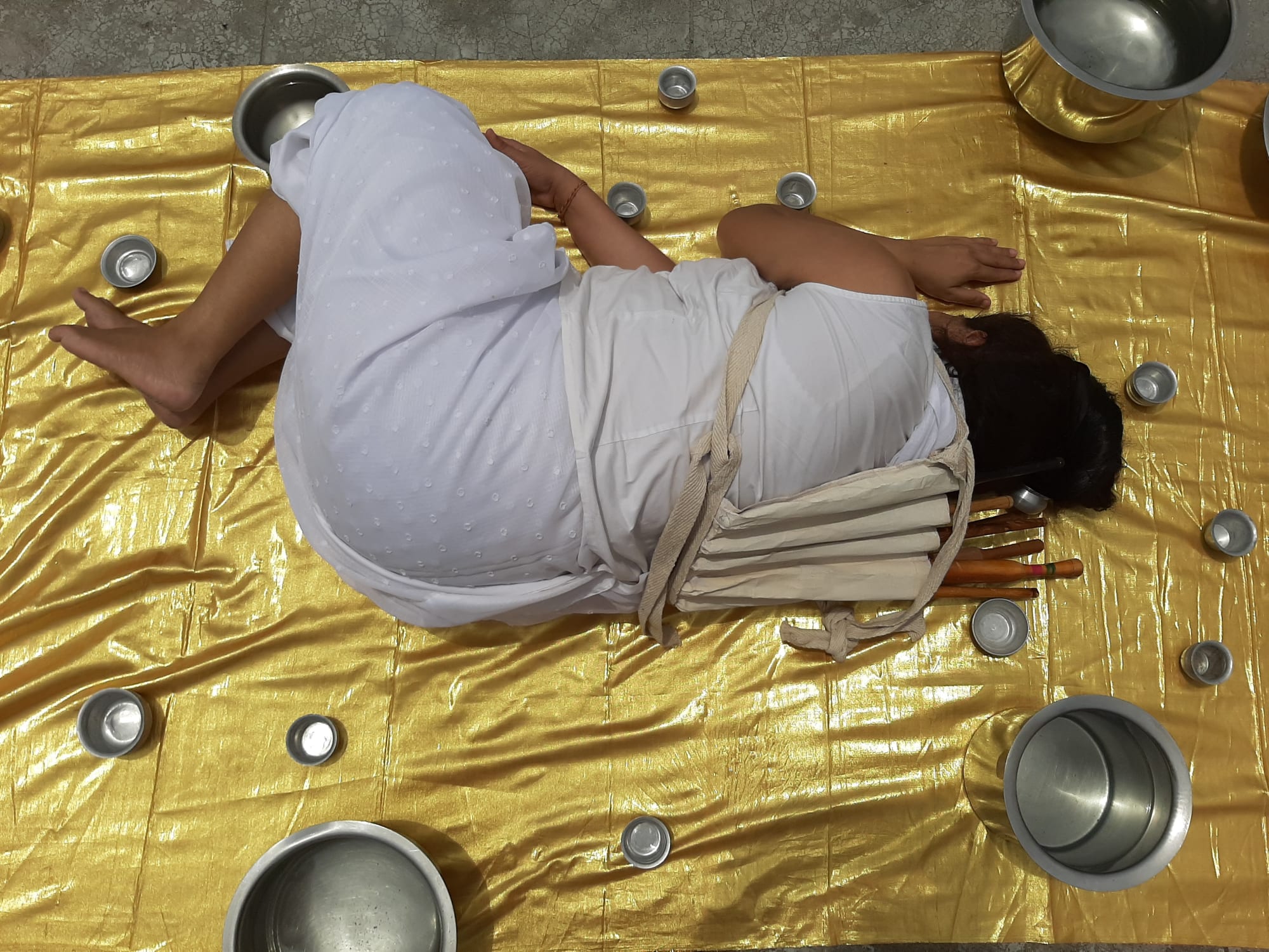 A digital photograph of a woman wearing white household clothes lying on a yellow golden sheet surrounded by steel utensils