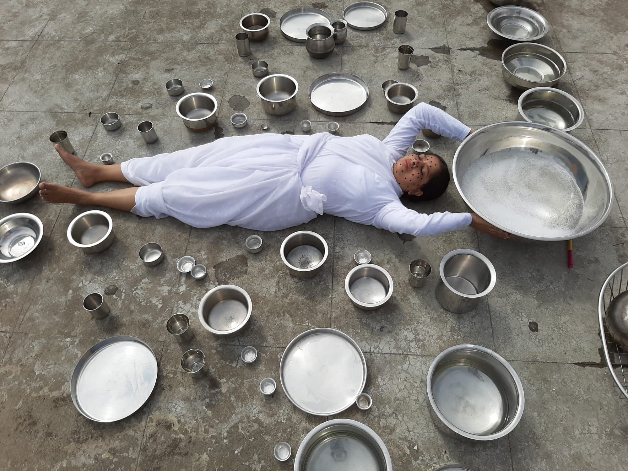 A digital photograph of a woman wearing white household clothes and lying on a cement floor surrounded by steel utensils