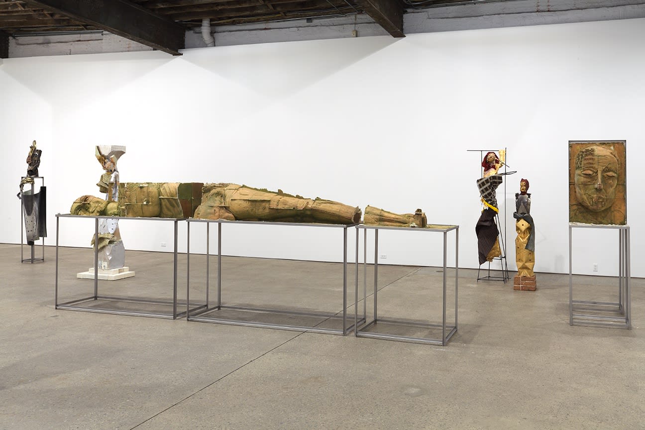 Installation view of Matthew Monahan's sixth solo show at Anton Kern Gallery, featuring sixteen sculptures, a set of drawings, two wall-mounted masks, and a single large figure in the back room. Long sculpture with other works.