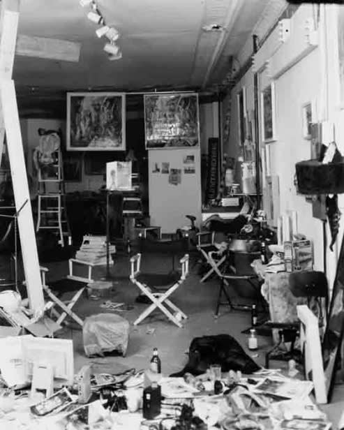 A black and white photo of a studio with director's chairs and many framed items hung on the wall, with detritus and clutter throughout